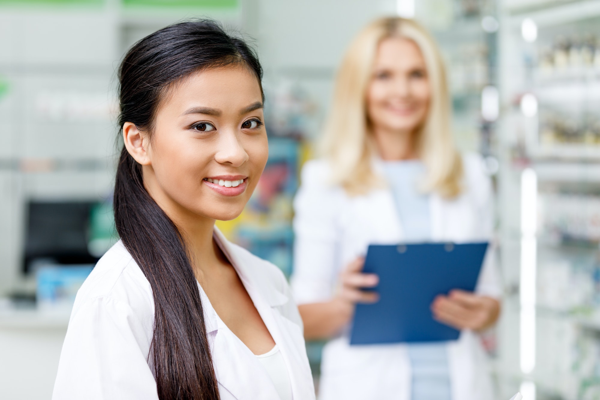 beautiful female asian pharmacist smiling at camera while colleague with clipboard standing behind